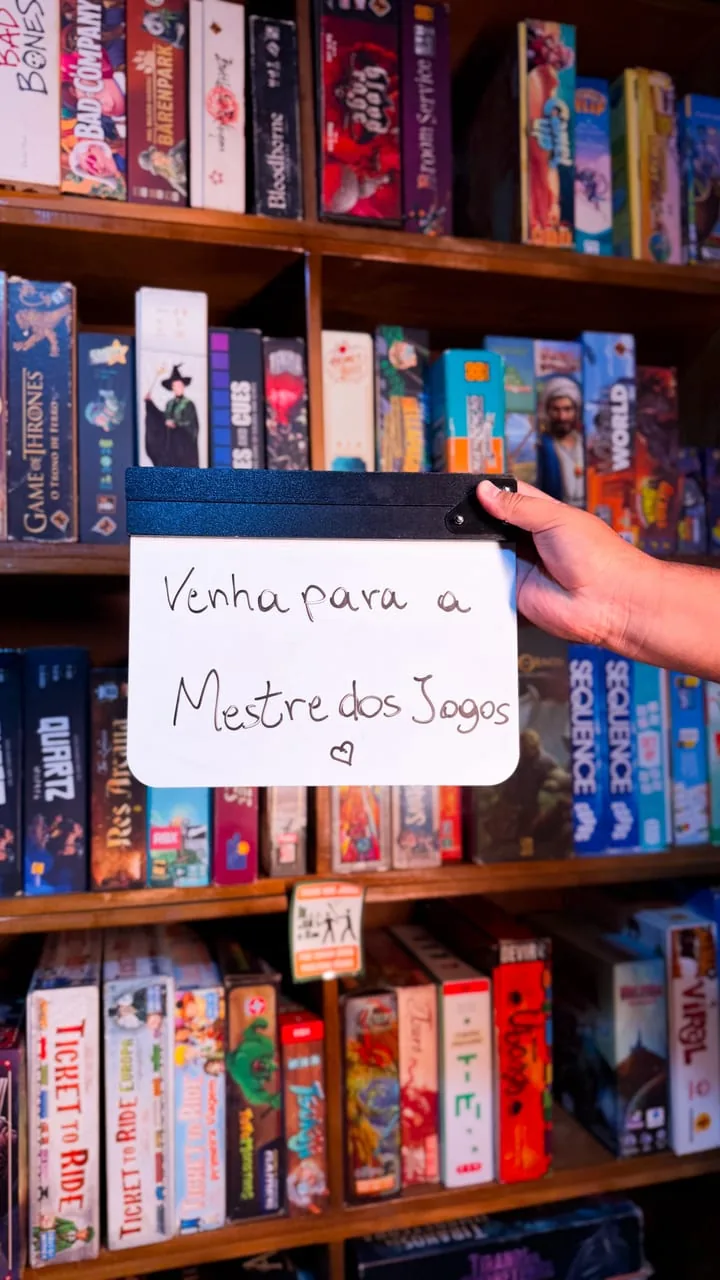Placa escrita à mão “Venha para a Mestre dos Jogos” com um coração, em frente à estante de jogos